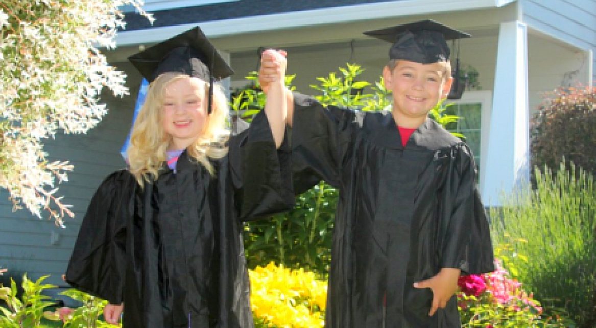 Two Little VIP kindergarten graduates in caps and gowns, hands raised, under the big tree